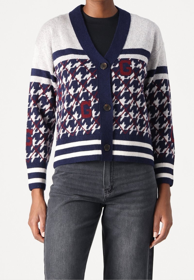 Personne portant un cardigan boutonné bleu marine, blanc et gris avec des motifs pied-de-poule et lettre G, associé à un jean gris foncé.