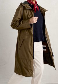 Olivgrön vattentät jacka med huva, dragkedjestängning och sidofickor, lager på lager över en marinblå ribbad tröja och beiga byxor.