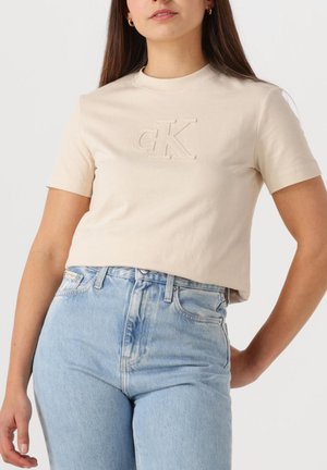 Mujer vistiendo una camiseta beige de manga corta con el logo "cK" en relieve y unos vaqueros de mezclilla azul claro de tiro alto, de pie con una mano en la cadera.