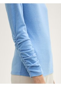 Top azzurro chiaro a maniche lunghe con maniche arricciate, texture morbida e scollatura rotonda, abbinato a un pantalone chiaro e neutro.