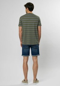Camiseta de rayas verdes con líneas blancas y marinas en un modelo, combinada con pantalones cortos de denim remangados y zapatos beige, vista desde atrás.