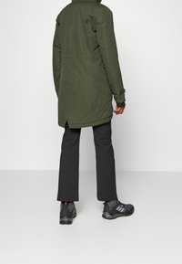 Cappotto lungo verde oliva con design dritto, tasche laterali e colletto regolabile, abbinato a pantaloni scuri e calzature nere.