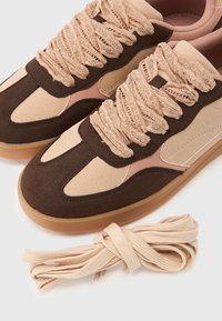 Ζευγάρι μπεζ και σκούρα καφέ σουέτ αθλητικά παπούτσια με παχιές κορδέλες κορδονιών, δίπλα σε ένα επιπλέον σετ μπεζ επίπεδων κορδονιών σε λευκό φόντο.