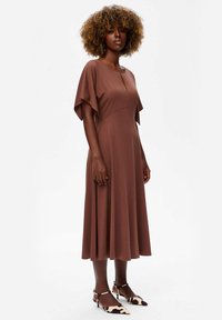 Vrouw met krullend haar, die ronde oorbellen draagt, een bruine midi-jurk met fladdermouwen en zwart-witte puntige hakken staat zijwaarts.