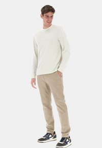 Camicia beige chiaro a maniche lunghe, color crema con logo, abbinata a pantaloni marrone chiaro e sneaker nere con accenti blu.
