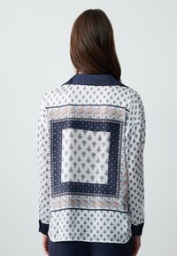 Blouse à manches longues en soie blanche avec des accents marine. Présente des motifs paisley complexes et un design bordé. Texture lisse et coupe décontractée.