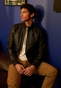 Veste bomber en cuir noir avec une fermeture éclair à l'avant, dotée de poches latérales et de poignets côtelés, portée sur un t-shirt blanc et un pantalon beige clair.