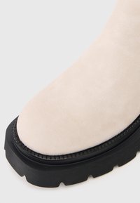 Zbliżenie na beżowy zamszowy czubek buta z grubą czarną gumową podeszwą oraz teksturowanym detalem na weltcie.