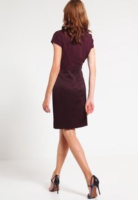 Robe bordeaux à manches courtes, corsage ajusté, jupe texturée et longueur genou. Portée avec des chaussures à talons hauts noires avec des accents bleus.