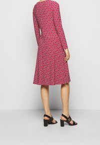 Czerwona sukienka z długimi rękawami, zdobiona geometrycznym wzorem w białym i niebieskim kolorze. Rozkloszowany dół, fakturowany materiał, w zestawie z czarnymi sandałami na obcasie.