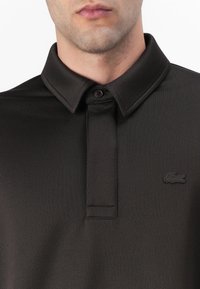Polo noir avec un tissu texturé, fermeture à boutons et col plat. Présente un logo noir discret sur la poitrine gauche.