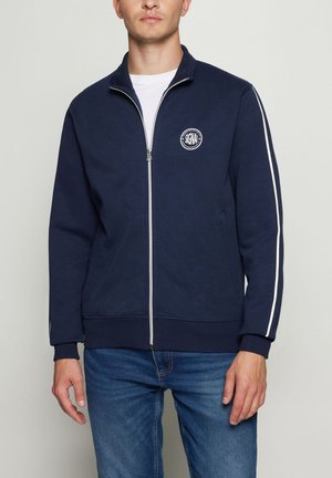 Mand iført navyblå lynlåstrøje med hvid stribe på ærmerne og cirkulært "SIGNAL" logo på venstre bryst, kombineret med blå jeans.