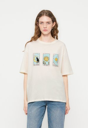 Ung kvinde iført en cremefarvet t-shirt med tarotkortdesigns mærket "Månen", "Solen" og "Stjernen", kombineret med blå jeans.