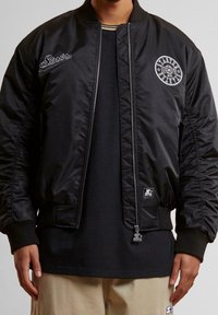 Veste bomber noire avec logo brodé, poignets côtelés, fermeture éclair et poches latérales. Extérieur lisse avec intérieur matelassé.