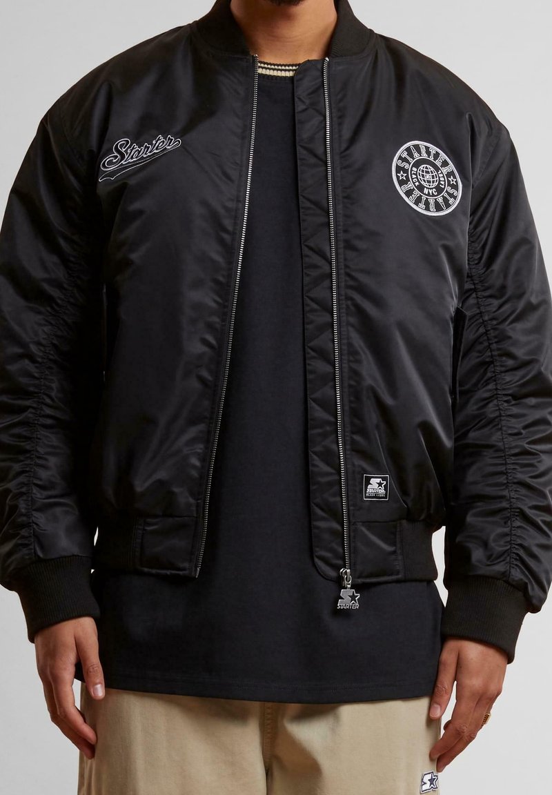 Veste bomber noire avec logo brodé, poignets côtelés, fermeture éclair et poches latérales. Extérieur lisse avec intérieur matelassé.