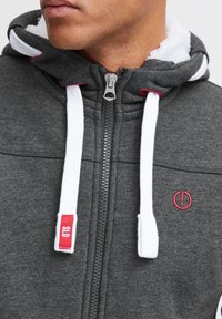 Grijze rits hoodie met een witte fleecelining, contrasterende rode accenten op de capuchon en schouder, en stevige witte koordjes.