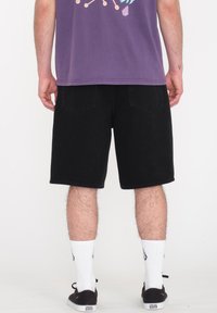 Shorts en denim noir avec poches arrière, coupe lâche et bords ourlés. Portés avec des chaussettes blanches et des baskets noires. Design simple, sans motifs visibles.