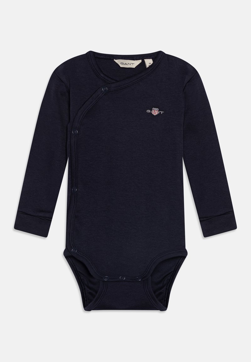 Jūras zila garām piedurknēm bodijs, kas izgatavots no mīksta auduma. Iezīmējas ar šķērsgriezuma dizainu un spiedpogu aizdari apakšā, kā arī ar piegrieztām piedurknēm. GANT logo priekšpusē.