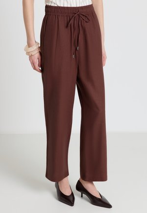 Donna che indossa pantaloni marroni a vita alta ampi con coulisse in vita, abbinati a décolleté marrone scuro con punta affilata e due braccialetti beige al polso sinistro.