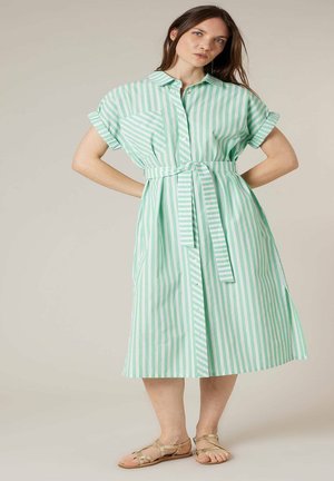 Femme debout, mains sur les hanches, portant une robe midi ceinturée à rayures verticales vertes et blanches et des sandales plates dorées.