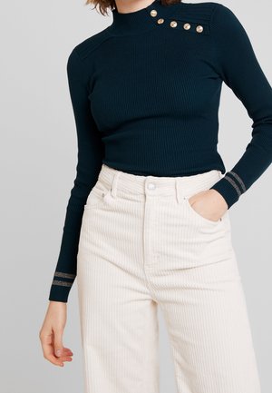 Top de manga larga en tono verde azulado oscuro con cuello alto y detalles de botones dorados en el hombro, combinado con pantalones de pana en color blanco roto.