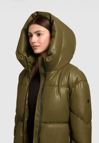 Olijfgroene pufferjack met een grote, gestructureerde capuchon, doorgestikte textuur en een rits aan de voorkant. Heeft een zwart accent op de mouw.