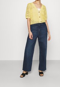 Gehaakte gele cardigan met korte pofmouwen, V-hals en houten knopen, gecombineerd met donkere blauwe wijde broek en zwarte open teensandalen.