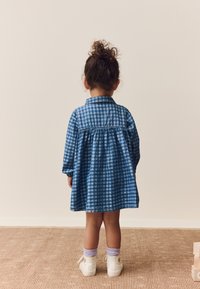 Mėlyna languota suknelė ilgomis rankovėmis ir apykakle, surinkta ties juosmeniu. Balti aukšti sportbačiai su šviesiai violetinėmis kojinėmis. Paprasta fonas.