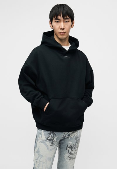 Must musta üle suurusega kapuutsiga dressipluus, millel on ees tasku, reguleeritava nööriga kapuuts ja minimalistlik bränding; kantud helehallide graafiliste teksadega.