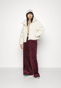 Veste en duvet créme coupée avec un col haut, associée à un pantalon large bordeaux. Casquette blanche avec un texte rose et des baskets accentuées de teal.