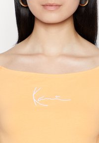 Πορτοκαλί τοπ με ανοιχτούς ώμους, φτιαγμένο από μαλακό ύφασμα, με λευκή κεντημένη υπογραφή στο στήθος, που αναδεικνύει τον χαλαρό σχεδιασμό.
