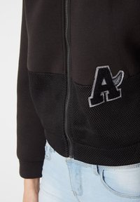 Chaqueta negra con cremallera que presenta un panel de malla texturizado en la parte inferior y un detalle bordado en gris de la letra "A" en el frente.