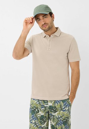 Beige Polohemd mit kurzen Ärmeln und drei Knöpfen, kombiniert mit einer grünen Kappe und bläulich-grünen Shorts mit Tropenblätter-Muster.