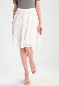 Weißer A-Linien-Rock aus Spitze mit kunstvollen, wirbelnden Mustern, strukturiertem Stoff und tailliertem Bund. Kombiniert mit einem grauen Oberteil und beigen High Heels.