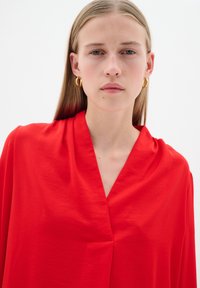 Blusa rossa senza maniche con scollatura a V e morbida texture, caratterizzata da una duttilezza minima e linee pulite. Orecchini a cerchio dorati come accenti.