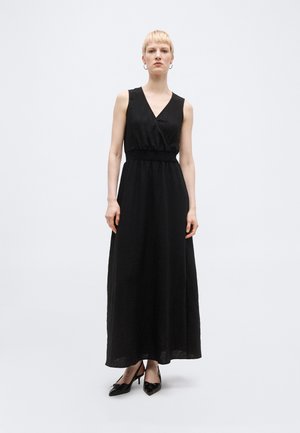 Frau mit kurzem blondem Haar, die ein ärmelloses schwarzes Maxikleid mit V-Ausschnitt und schwarzen spitzen Schuhen trägt, vor einem einfarbigen Hintergrund stehend.