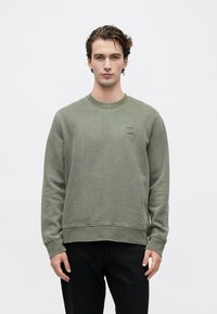 Mlad moški s kratkimi valovitimi lasmi, oblečen v olivno zeleno jopico in črne hlače, ki stoji ob ozadju v beli barvi.