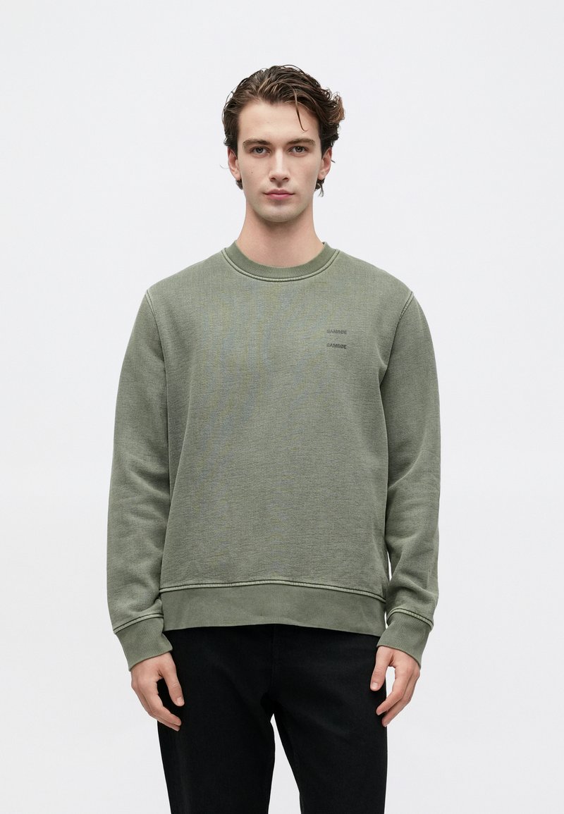 Mlad moški s kratkimi valovitimi lasmi, oblečen v olivno zeleno jopico in črne hlače, ki stoji ob ozadju v beli barvi.