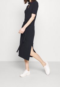 Gestricktes, kurzärmliges Kleid in Marineblau mit Kragen und seitlichen Schlitzen; kombiniert mit weißen Sneakers. Verfügt über einen Taillengürtel und subtile Designdetails.