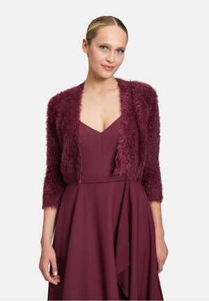 Cardigan court en laine moelleuse de couleur bourgogne avec des manches trois-quarts ; porté par-dessus une robe à décolleté en V profond assortie. Surface texturée et matériau doux.