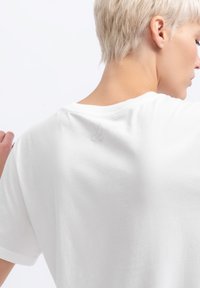 Witte katoenen t-shirt met een achteraanzicht, een ronde hals, korte mouwen en een subtiele schaarlogo in lichtgrijs gedrukt.