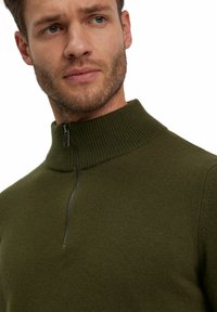 Maglione verde lavorato a maglia con colletto alto e chiusura a mezza zip. Presenta una finitura testurizzata e un design aderente.