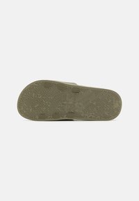 Khaki gummisål med teksturert gripe mønster, seks runde knotter for grep, og et sentralt logoavtrykk. Flat og svakt buet form.