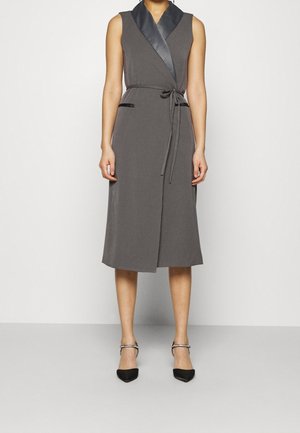 Szara, bezrękawna sukienka kopertowa do kolan z czarnym satynowym kołnierzem, wiązanym paskiem w talii oraz czarnymi, spiczastymi szpilkami z paskami wokół kostki.