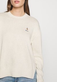 Beige gestrickter Pullover mit Rundhalsausschnitt und gerippten Bündchen, mit einem kleinen gestickten Logo auf der Brust. Weiche Textur, lässige Passform.