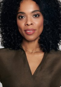 Brązowa, fakturowana bluzka z dekoltem w kształcie V. Nosi delikatny naszyjnik ze złotego łańcuszka z małymi, nieregularnymi ogniwami. Włosy kręcone, sięgające ramion.