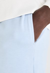 Lyseblå joggebukser med en glatt tekstur som har en sidelomme. Livsnormen er laget av hvit ribbestrikket stoff som gir kontrast.
