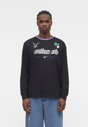 Mężczyzna z zaplecionymi włosami, ubrany w czarną, długą koszulkę Nike SB z logo i niebieskie dżinsy, stojący na białym tle.