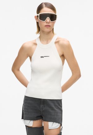 Donna che indossa occhiali da sole con montatura bianca oversize, top bianco a collo alto con scritta e pantaloncini di jeans neri con bordi sfrangiati su sfondo bianco.