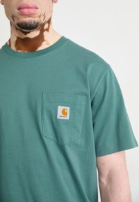 Teal cotton t-shirt with a crew neckline and a chest pocket featuring a white patch with a stylized orange logo.